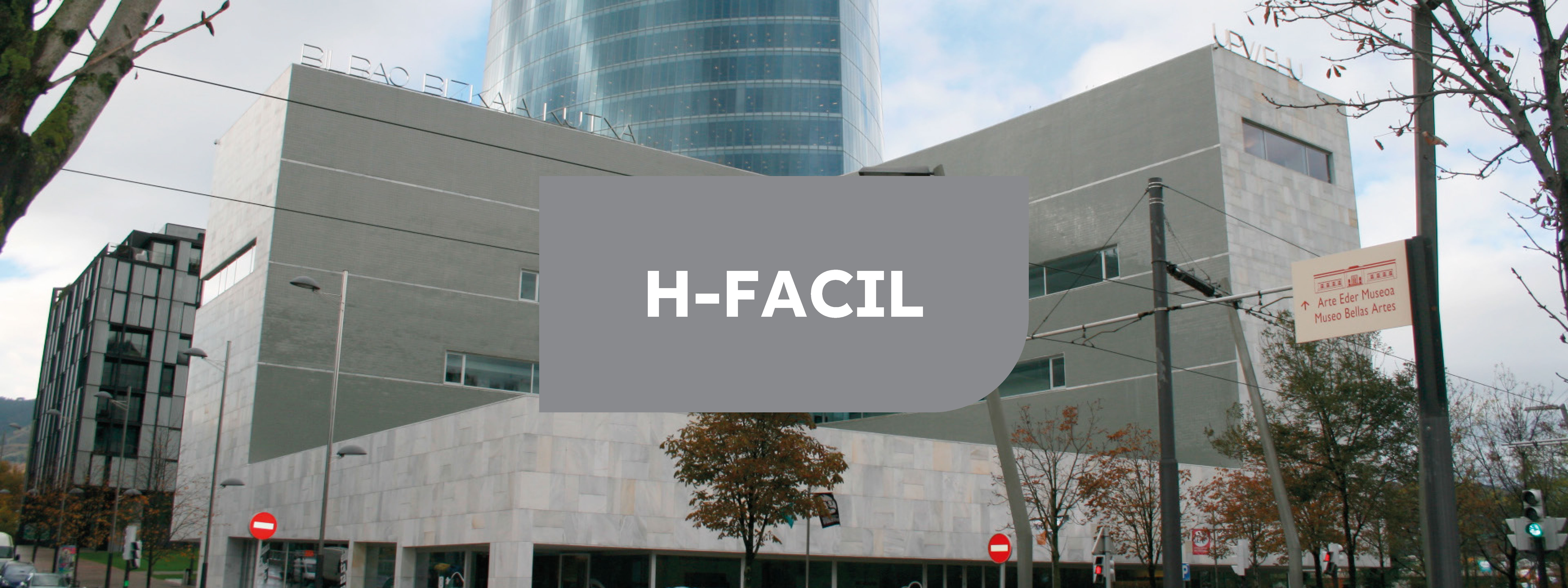 Edificio moderno gris en forma de "L" con un cartel en el centro con fondo gris y letras blancas "H-FACIL"