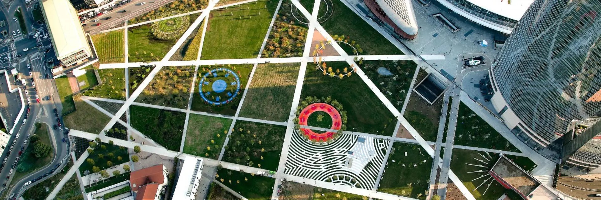 a vista de pájaro: parque con senderos lineales blancos y varias circunferencias, rodeado de edificios futuristas.