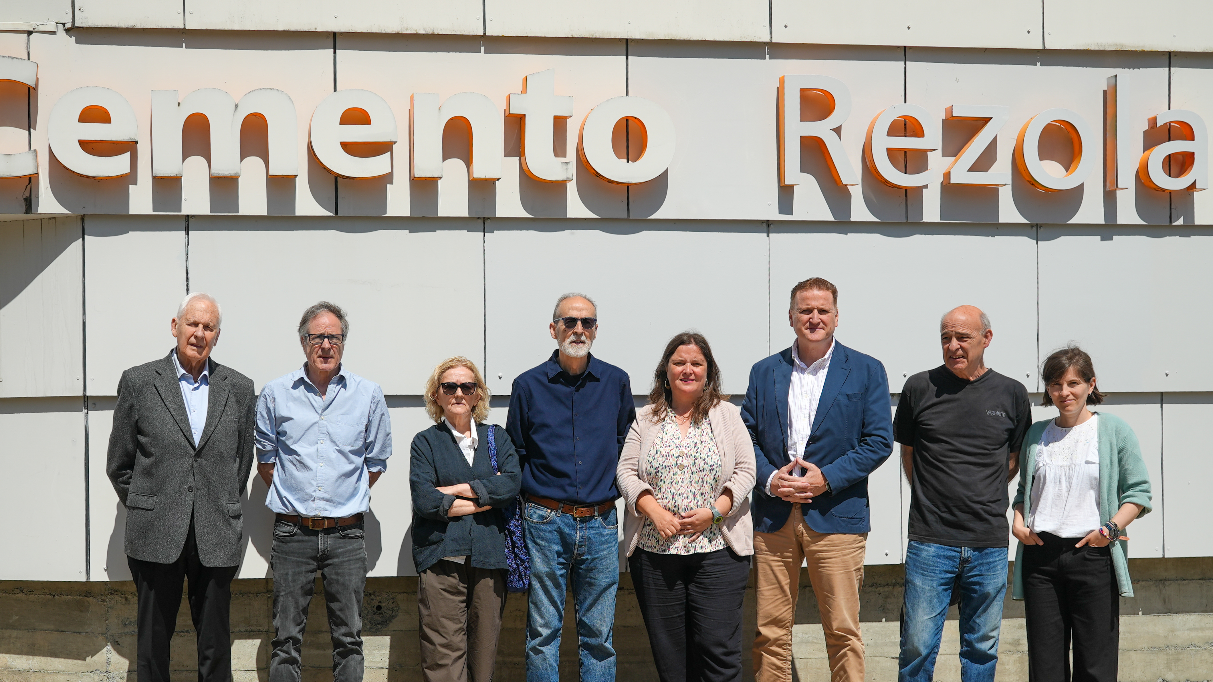 Un grupo de ocho personas posa al aire libre frente a un edificio con el letrero "Cemento Rezola" en la parte superior. Todos están de pie, en una fila, con expresiones neutras o sonrientes. El fondo muestra una pared blanca estructurada en paneles. La luz solar ilumina la escena.