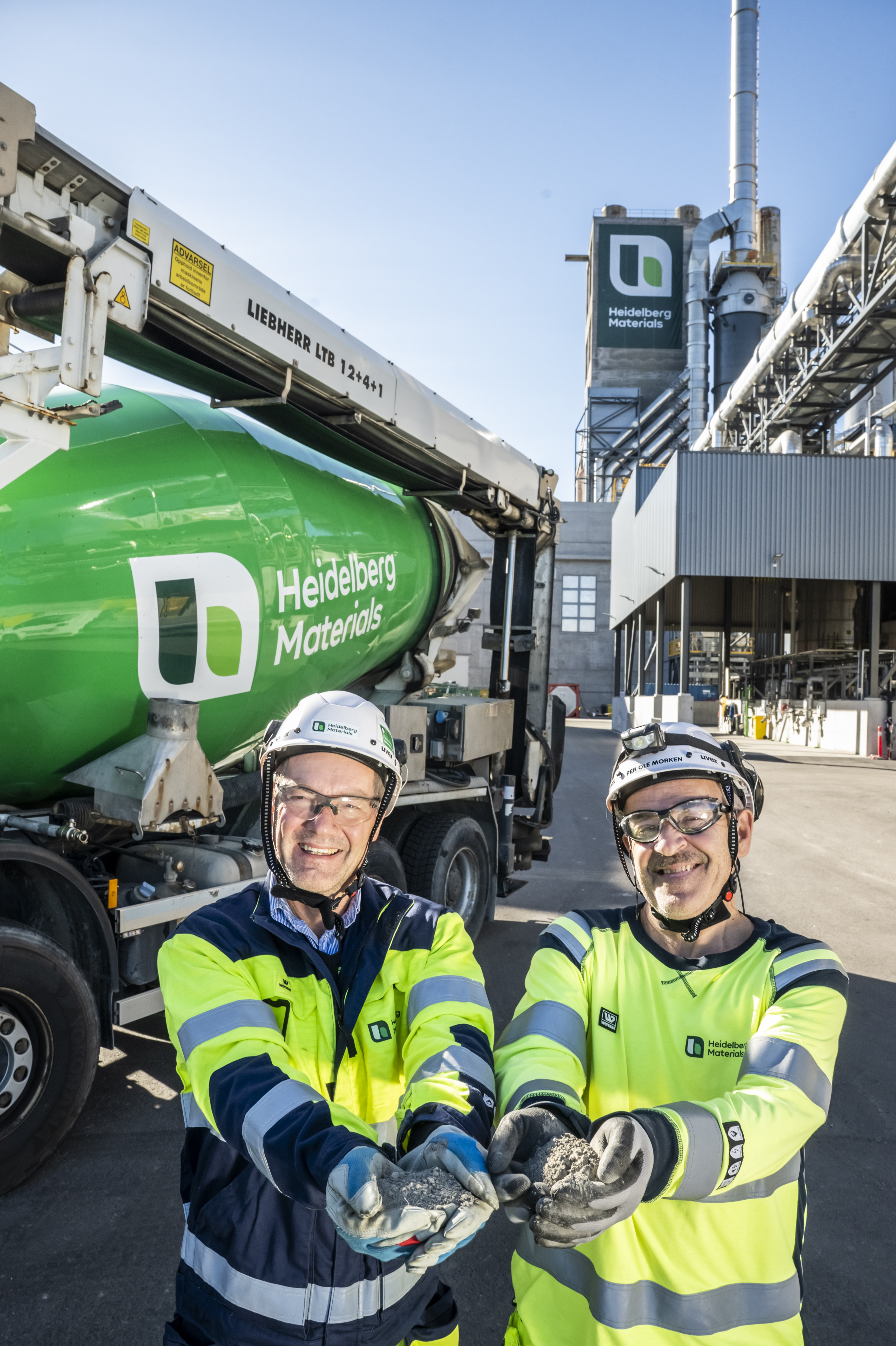 Dos trabajadores con equipo de seguridad sostienen material en sus manos frente a una planta industrial y un camión mezclador verde con el logotipo de Heidelberg Materials. Al fondo, estructuras industriales y una torre con el logo de la empresa visible.