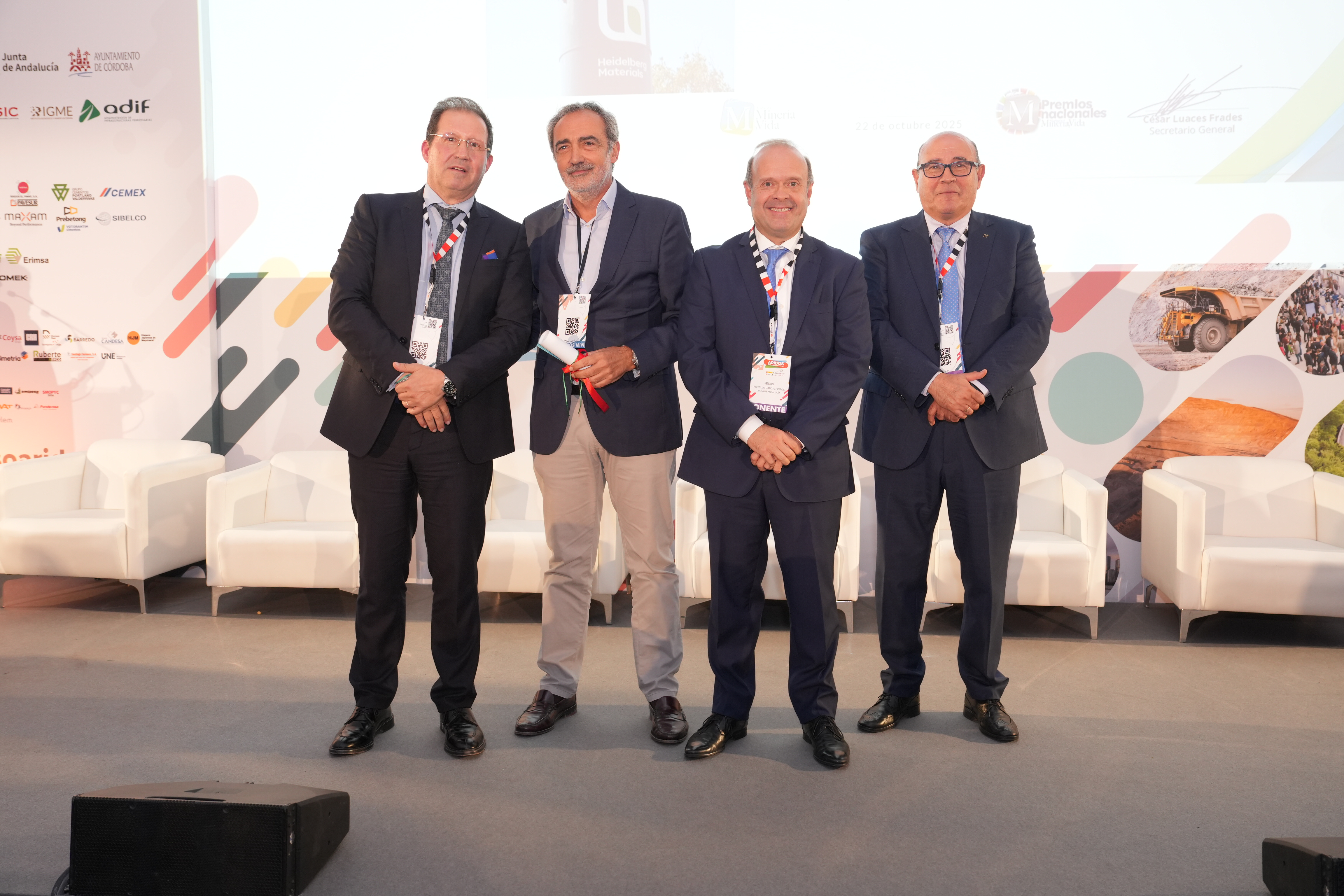 Cuatro hombres vestidos formalmente posan juntos en un escenario durante un evento. Detrás de ellos se observan gráficos coloridos, logotipos de patrocinadores y sillones blancos alineados.