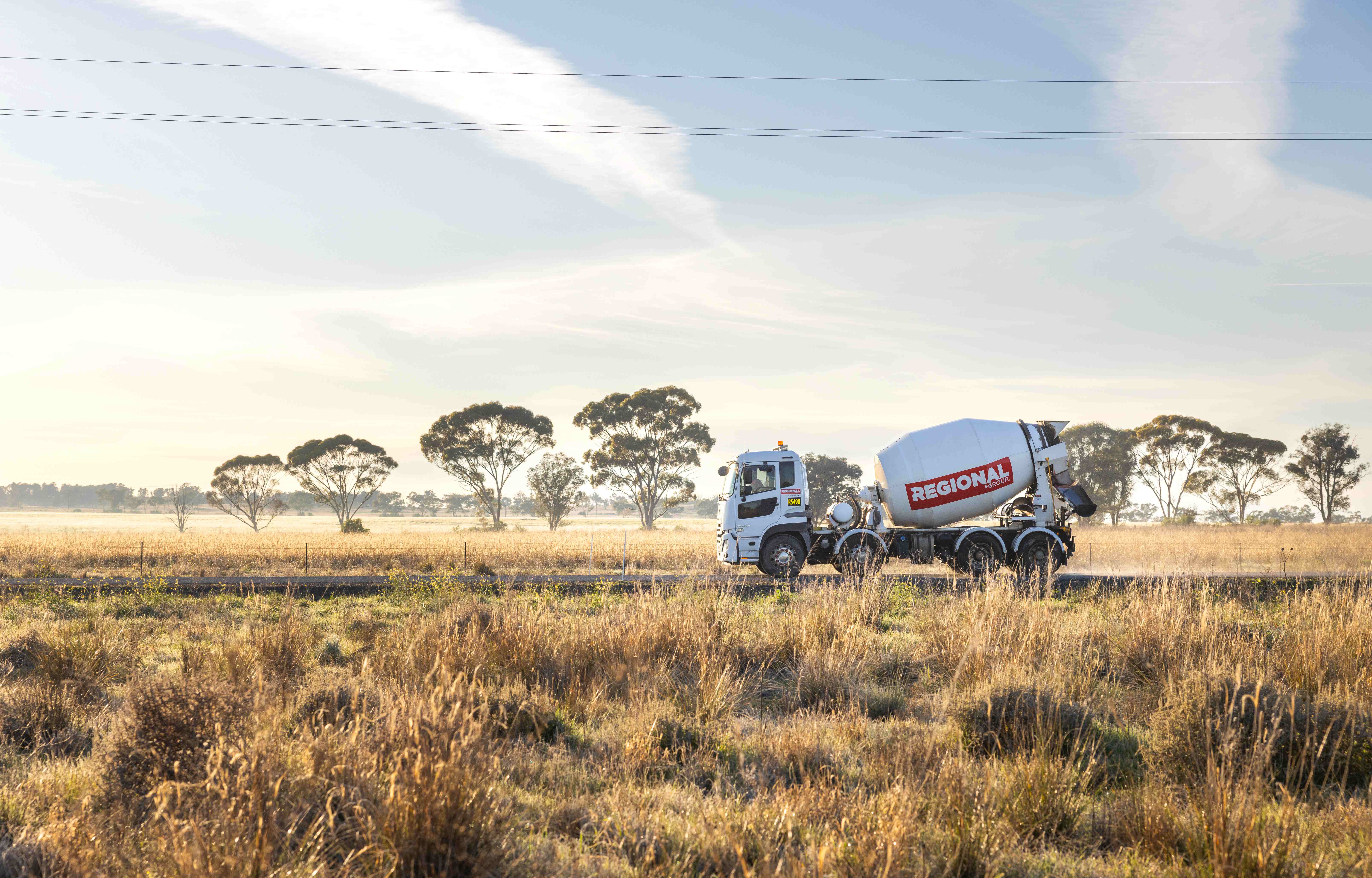 Camión mezclador de cemento estacionado en un campo abierto bajo un cielo despejado, con árboles dispersos en el fondo y pastizales en primer plano.