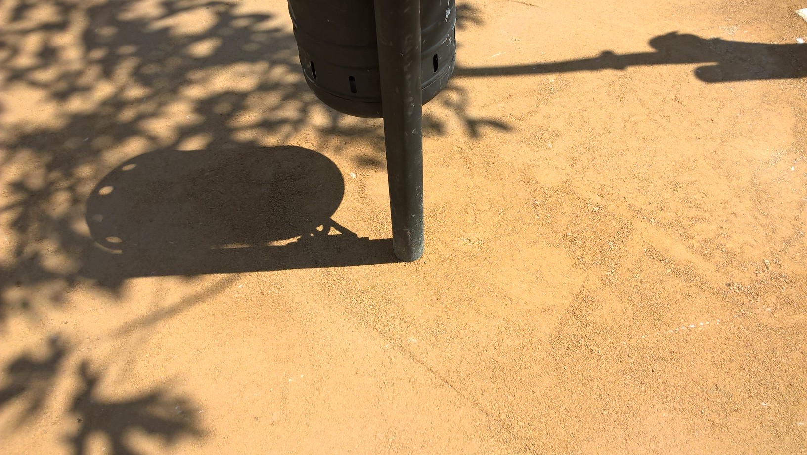 Detalle de suelo de tierra compacta en parque con sombra de hojas y pie de papelera