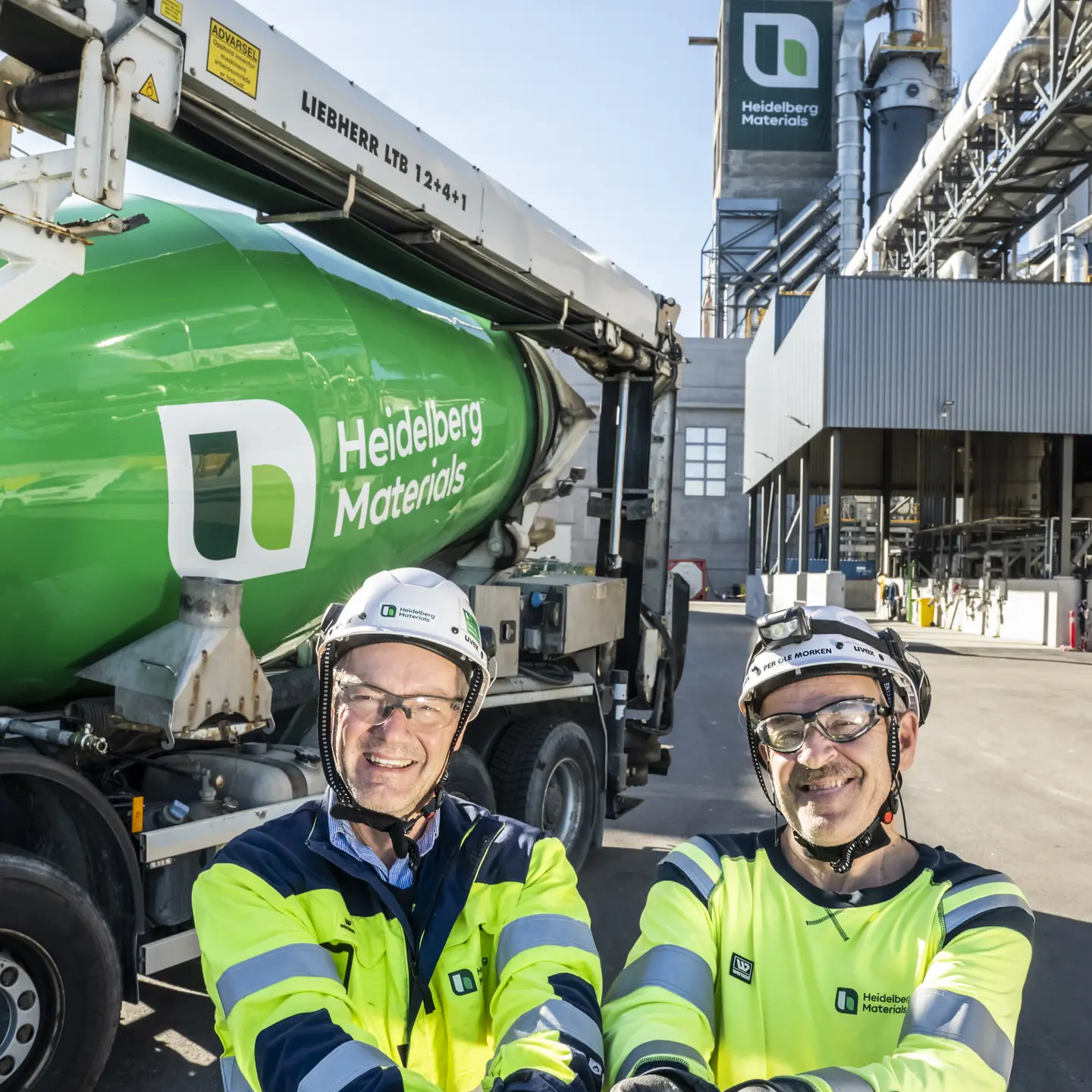 Dos trabajadores con equipo de seguridad sostienen material en sus manos frente a una planta industrial y un camión mezclador verde con el logotipo de Heidelberg Materials. Al fondo, estructuras industriales y una torre con el logo de la empresa visible.