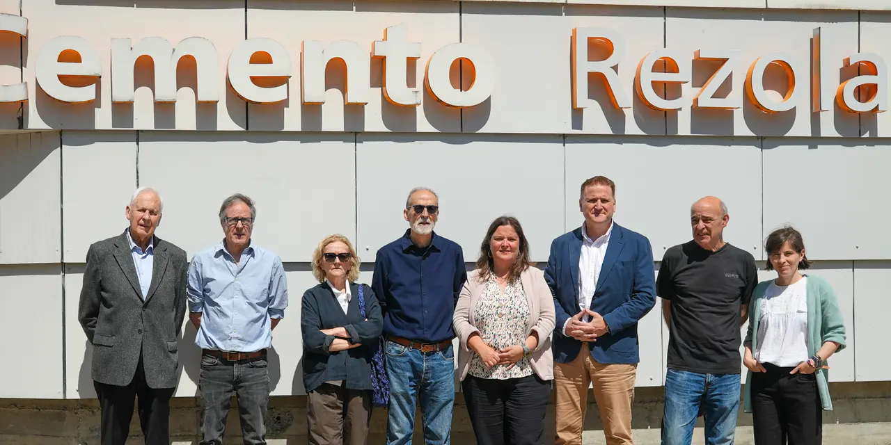 Un grupo de ocho personas posa al aire libre frente a un edificio con el letrero "Cemento Rezola" en la parte superior. Todos están de pie, en una fila, con expresiones neutras o sonrientes. El fondo muestra una pared blanca estructurada en paneles. La luz solar ilumina la escena.