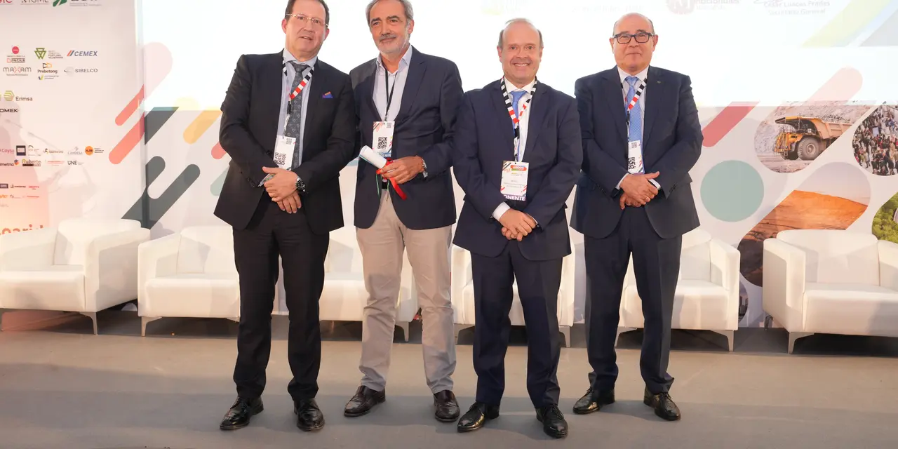 Cuatro hombres vestidos formalmente posan juntos en un escenario durante un evento. Detrás de ellos se observan gráficos coloridos, logotipos de patrocinadores y sillones blancos alineados.