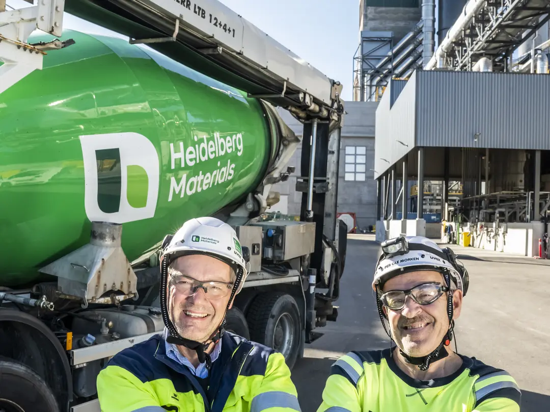 Dos trabajadores con equipo de seguridad sostienen material en sus manos frente a una planta industrial y un camión mezclador verde con el logotipo de Heidelberg Materials. Al fondo, estructuras industriales y una torre con el logo de la empresa visible.
